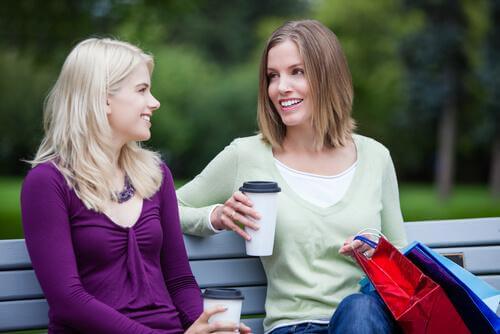 To unge kvinner sitter på en benk og drikker kaffe og snakker med hverandre