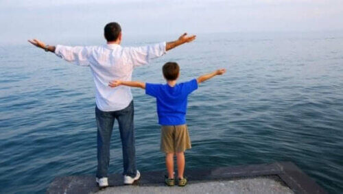 Far og sønn med utstrakte armer foran vann.