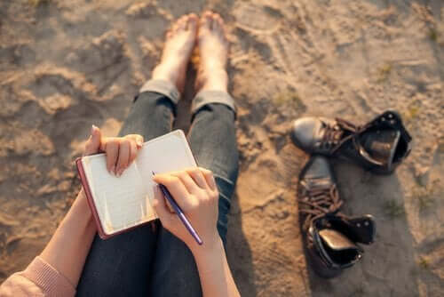 Kvinne sitter barfot på en strand og skriver dagbok