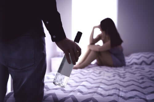 En kvinne sitter på sengen, mens en mann står med en vinflaske i hånden står litt lengre vekk.
